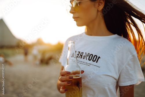 Sticker Young girl drinking beer from a bottle on the beach at sunset during  summer vacation