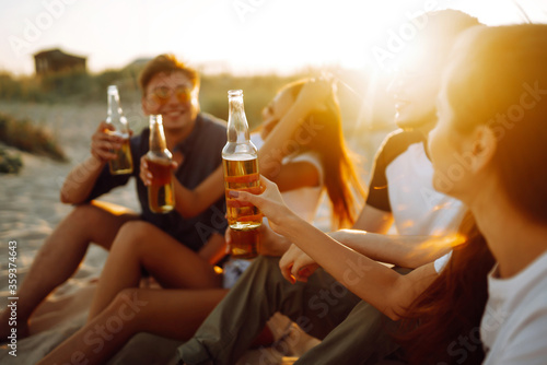 Young people sitting together at beach, drinking beer and having a party. Group of friends cheers with beers at the beach during summer vacation.