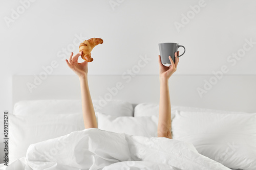 morning, comfort and people concept - young woman with cup of coffee and croissant lying in bed at home bedroom
