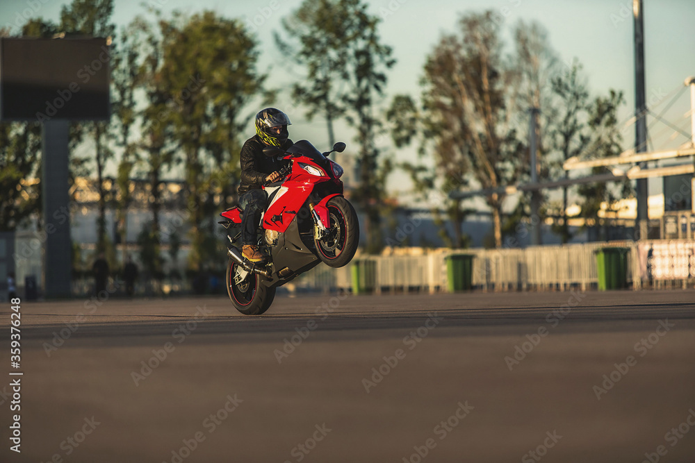 Stunt rider in helmet and black leather jacket riding red sports motorbike on one wheel