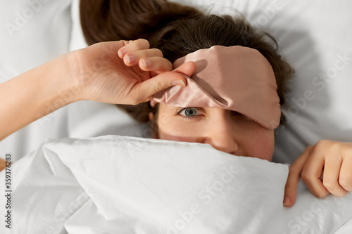 people, bedtime and rest concept - happy woman in bed with eye sleeping mask peeking from under blanket