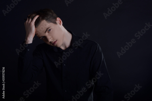 Studio portrait of a guy in a black shirt on a black background. A handsome young guy of European appearance. The guy cares for himself, touches his hair, haircuts and gels for men's hair
