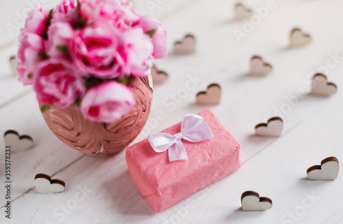 Vintage background with hearts, gift box and bouquet of pink roses in vase on white wooden board.