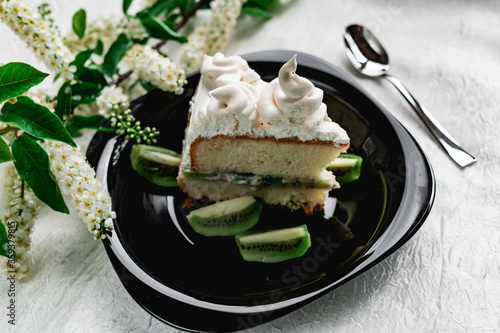 A slice of cake with meringue cream on a glossy black plate with slices of fresh kiwi. Cake decorated with a sprig of fragrant cherry blossoms