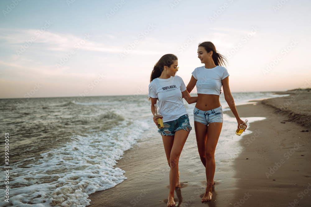 Young girls walking on the beach and drink beers at sunset, during summer vacation. Holidays, relax and lifestyle concept. Young girls walking on the beach and drink beers at sunset, during summer vacation. Holidays, relax and lifestyle concept.