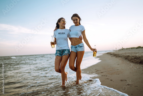 Young girls walking on the beach and drink beers at sunset, during summer vacation. Holidays, relax and lifestyle concept.