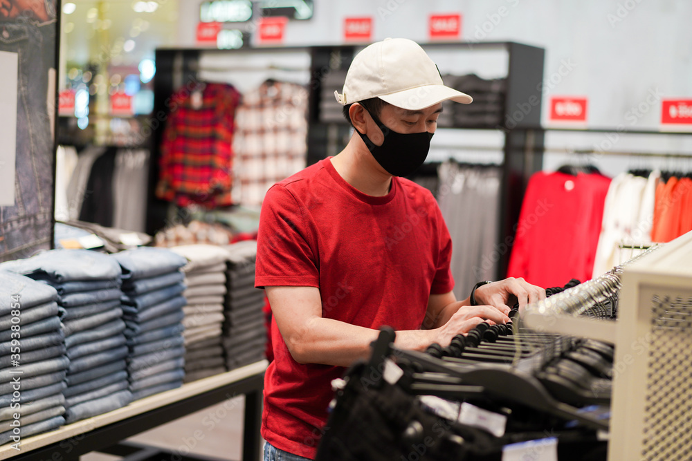 Young Asian man with a white cap and black protective mask on face shopping for clothes in shopping mall. Coronavirus protection and air pollution protection. Young Asian man with a white cap and black protective mask on face shopping for clothes in shopping mall. Coronavirus protection and air pollution protection.