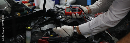 Close-up view of open trunk and male mechanic using multimeter or voltmeter checking a car battery level. Professional restoration workshop and repair concept