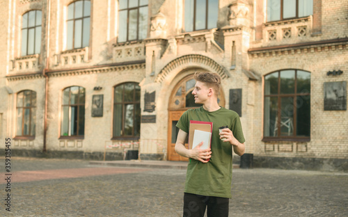 Handsome young male stands with books and a cup of coffee in his hands on a university building background and looks away with a smile on face. Positive guy in casual clothes drinking coffee at break. © bodnarphoto