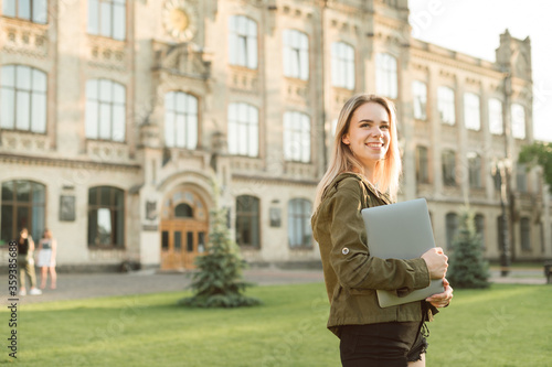 Portrait of happy lady in casual clothes standing on grass in campus on background of university building with laptop in hands, looking away at copy space and smiling.Girl is walking college yard