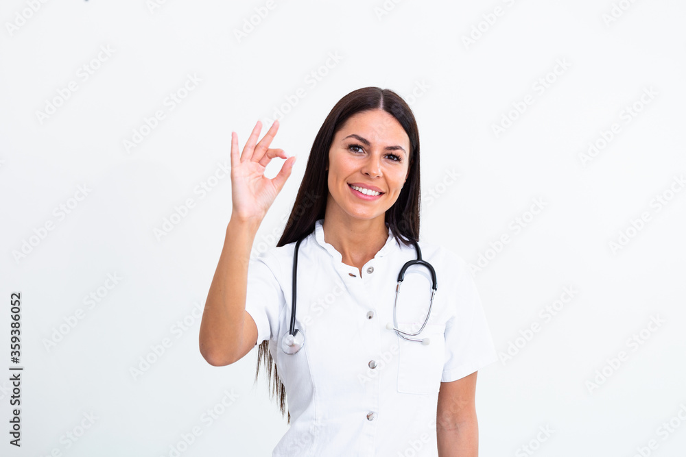 Medical concept of young beautiful female doctor in white coat with phonendoscope showing ok sign. Woman hospital worker looking at camera and smiling, studio, white background Medical concept of young beautiful female doctor in white coat with phonendoscope showing ok sign. Woman hospital worker looking at camera and smiling, studio, white background