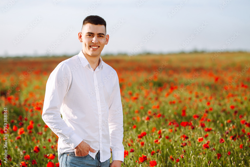 Portrait of a young man standing in a poppy field. Smiling boy being confident and feeling freedom among red poppy flowers on the sunset. Love, family value and peace concept.