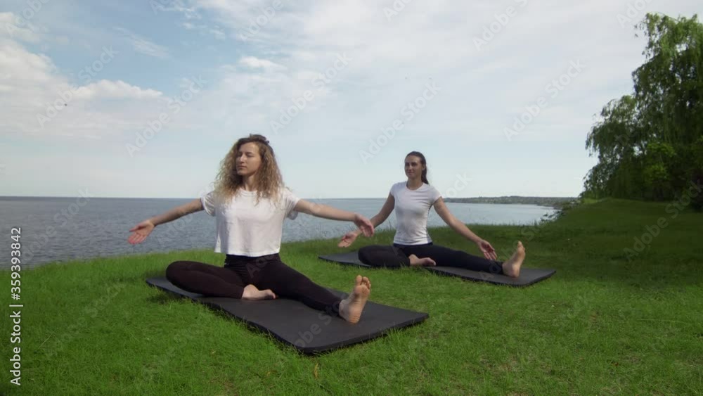 Two women meditating outdoors seated on the edge of coast near the sea, half lotus pose folded fingers mudra gesture do yoga exercise in morning. Two women practicing Revolved Head to Knee pose Two women meditating outdoors seated on the edge of coast near the sea, half lotus pose folded fingers mudra gesture do yoga exercise in morning. Two women practicing Revolved Head to Knee pose