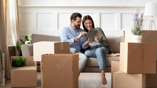 Excited young couple having fun with tablet on moving day, looking at mobile device screen, resting on couch in living room with cardboard boxes with belongings, relocation and mortgage concept © fizkes