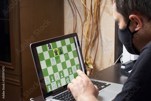 A man in a medical mask solves a chess problem online. Chess board with figures on a laptop screen. Leisure in self-isolation, in the twilight. International chess day. photo