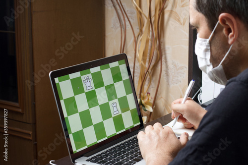 A man in a medical mask solves a chess problem online. Chess board with figures on a laptop screen. Leisure in self-isolation. International chess day. photo