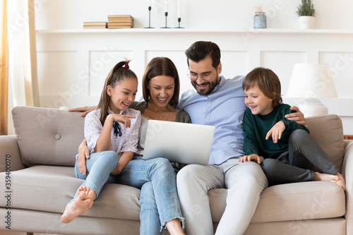 Happy family with children relaxing on couch with laptop, smiling young mother and father hugging adorable son and daughter, looking at computer screen, shopping, watching movie online