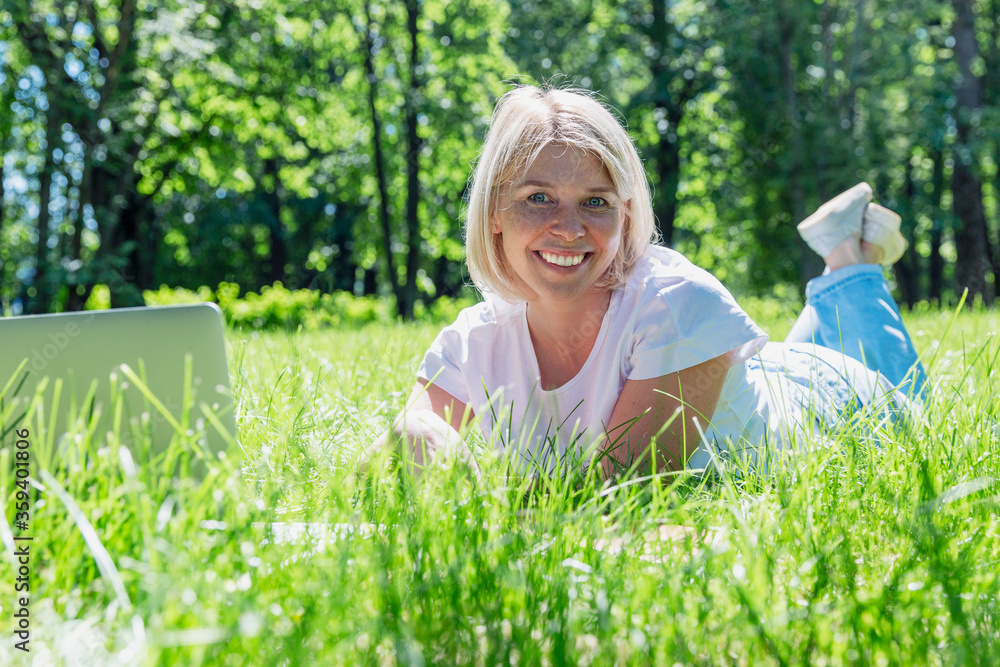 A smiling young blonde woman lies on the grass in a park with a laptop on a summer sunny day. Blogging, online communication, distance work and training. A smiling young blonde woman lies on the grass in a park with a laptop on a summer sunny day. Blogging, online communication, distance work and training.