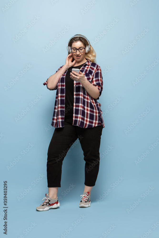 Young woman in casual wear with smartphone and headphones on blue background. Bodypositive character, feminism, loving herself, beauty concept. Plus size beautiful businesswoman. Inclusion, diversity.