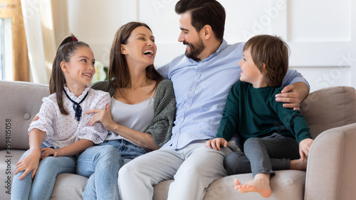 Happy mother and father hugging with children, embracing, laughing, sitting on couch, family having fun at home, parents with adorable son and daughter spending leisure time on weekend together