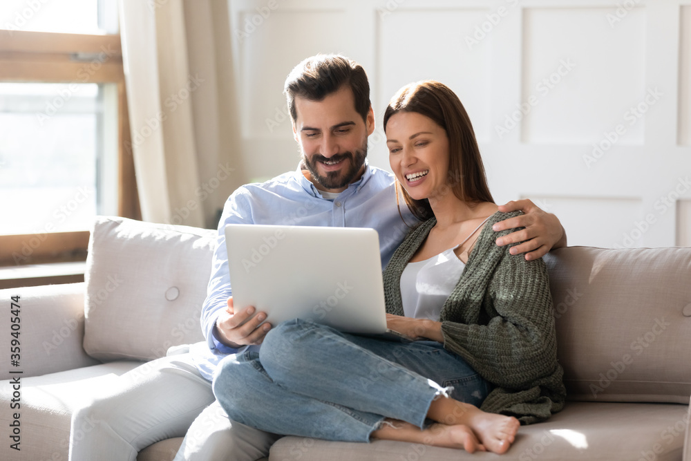Fototapeta premium Happy young couple using laptop together, looking at screen, watching movie or video, shopping or chatting online, smiling man and woman relaxing on cozy sofa, having fun with electronic device