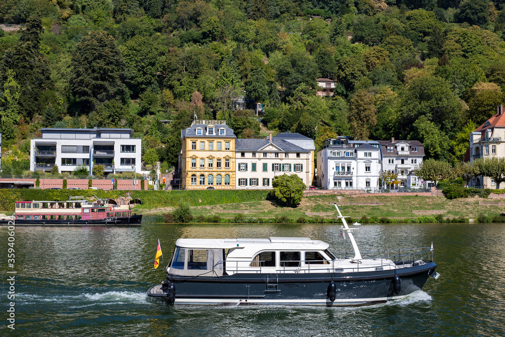 View of residential buildings. Houses on the river bank. A boat floats on the water. There are many beautiful green trees around. Modern area near the historic city of Heidelberg in Germany. View of residential buildings. Houses on the river bank. A boat floats on the water. There are many beautiful green trees around. Modern area near the historic city of Heidelberg in Germany.