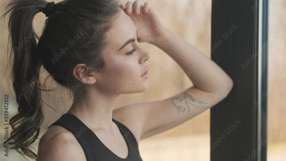 A close-up view of a charming pretty young woman in a black tracksuit is looking to the side standing near window in a gray studio A close-up view of a charming pretty young woman in a black tracksuit is looking to the side standing near window in a gray studio