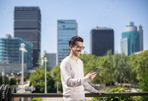 Joven trabajador con bigote y gafas sonriente mirando el teléfono móvil con un fondo de rascacielos a su espalda