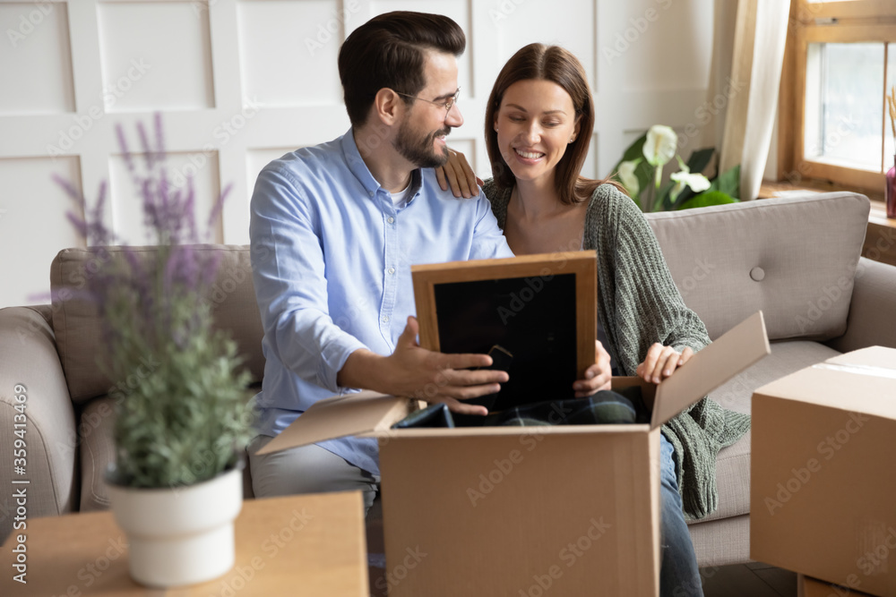 Happy wife and husband looking at photo frame, sharing memories, sitting on couch in living room, young couple unpacking cardboard boxes with belongings on moving day, relocation and mortgage Happy wife and husband looking at photo frame, sharing memories, sitting on couch in living room, young couple unpacking cardboard boxes with belongings on moving day, relocation and mortgage