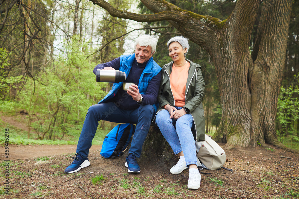 Full length portrait of cheerful senior couple enjoying hot tea from flask while resting during hike in autumn forest, copy space Full length portrait of cheerful senior couple enjoying hot tea from flask while resting during hike in autumn forest, copy space