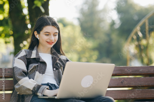 Caucasian woman in headset typing on the laptop computer on the bench in the park. Female student makes conference video call on laptop computer talks with web tutor, online teacher in webcam chat