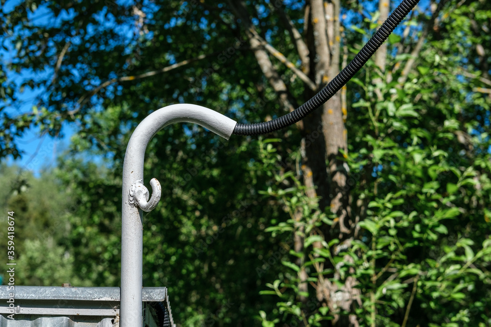 An electric wire in a black corrugation is connected to the house through a metal pipe bent down, against the background of trees in the garden and blue sky. An electric wire in a black corrugation is connected to the house through a metal pipe bent down, against the background of trees in the garden and blue sky.