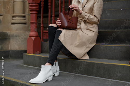 Fashionable young woman in black jeans, beige trench coat and white cowboy boots with brown handbag sitting on the steps .Outdoor . Fashion. Stylish .