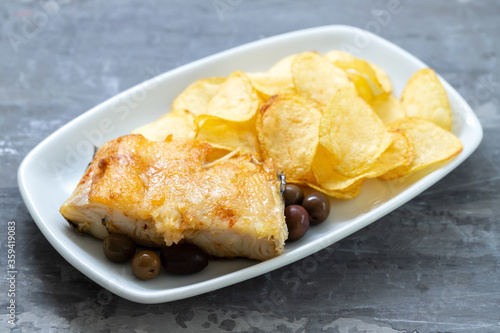 fried cod fish with olives and potato chips on white dish on ceramic background