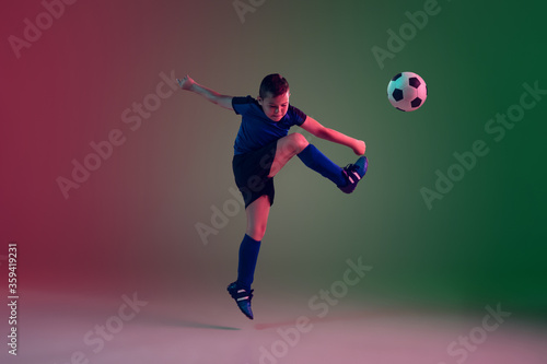 Teen male football or soccer player on gradient background in neon light. Caucasian expressive boy training  practicing on the run  in jump. Concept of sport  competition  winning  motion  action.