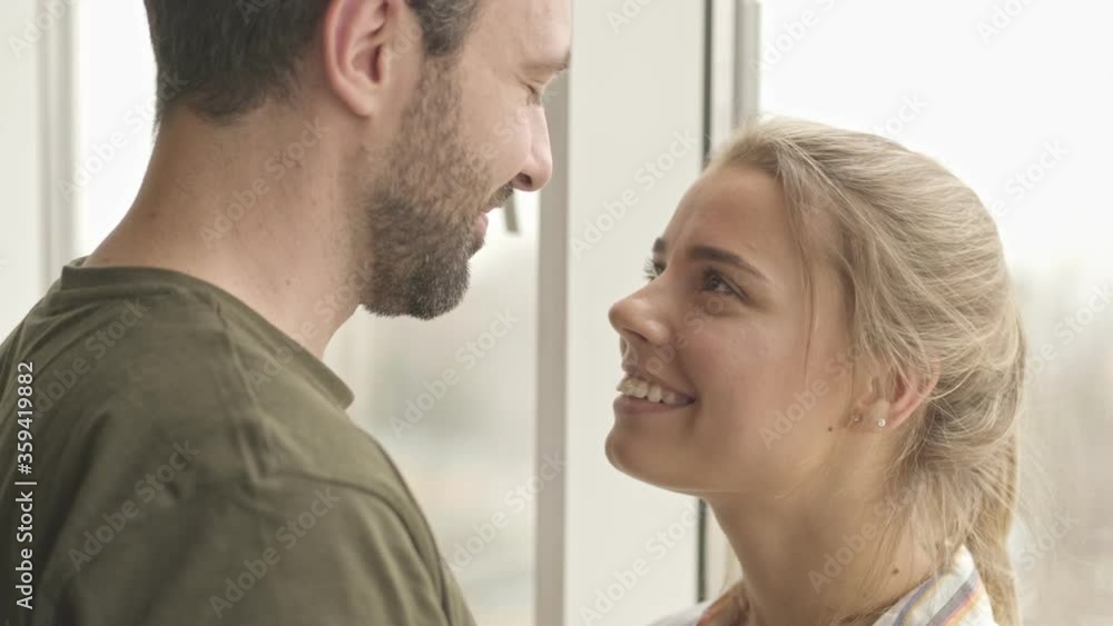 A lovely young couple are looking to each other and hugging while standing near window at home A lovely young couple are looking to each other and hugging while standing near window at home