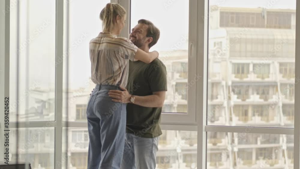 An attractive young couple man and woman are hugging and kissing while standing near window at home An attractive young couple man and woman are hugging and kissing while standing near window at home