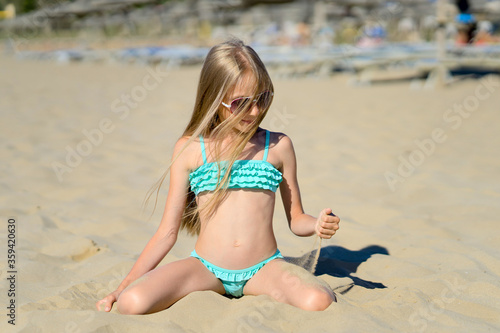 A girl with long blonde hair and in sunglasses sits on the beach and sprinkles sand on the body