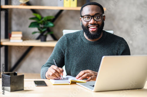 Cheerful African-American male student or worker is watching online lectures or webinars and writing notes in a notebook. A black guy sits at the desk with laptop, looks at camera and smiles
