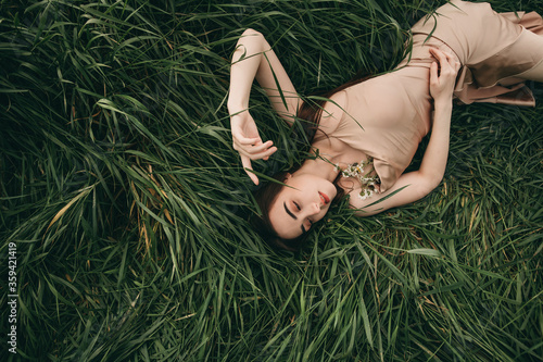 Young beautiful woman with long brunette hair in a beige dress lies on the green grass. Summer, stylish, nature, rest.