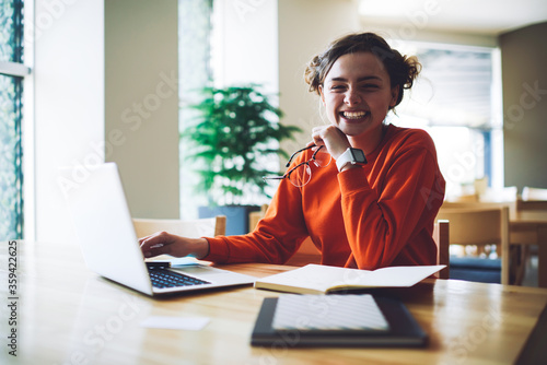 Half length portrait of emotional cute hipster girl sitting in college and preparing for exams while looking at camera and laughing.Cheerful teen with eyeglasses in hand smiling
