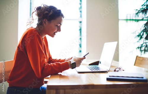 Smiling teenager browsing internet websites and doing shopping online on digital smartphone sitting at laptop and books in college interior.Young woman in good mood chatting with friends via cellular photo