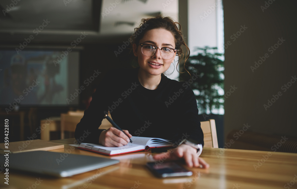 Portrait of intelligent student with optical spectacles writing some records in textbook while sitting at table with laptop computer in coworking space and looking at camera during exam preparation Portrait of intelligent student with optical spectacles writing some records in textbook while sitting at table with laptop computer in coworking space and looking at camera during exam preparation