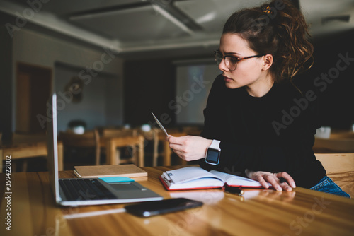 Pensive female entrepreneur in trendy spectacles for better vision sitting at modern laptop device and textbooks in coworking space while looking at business card reading information