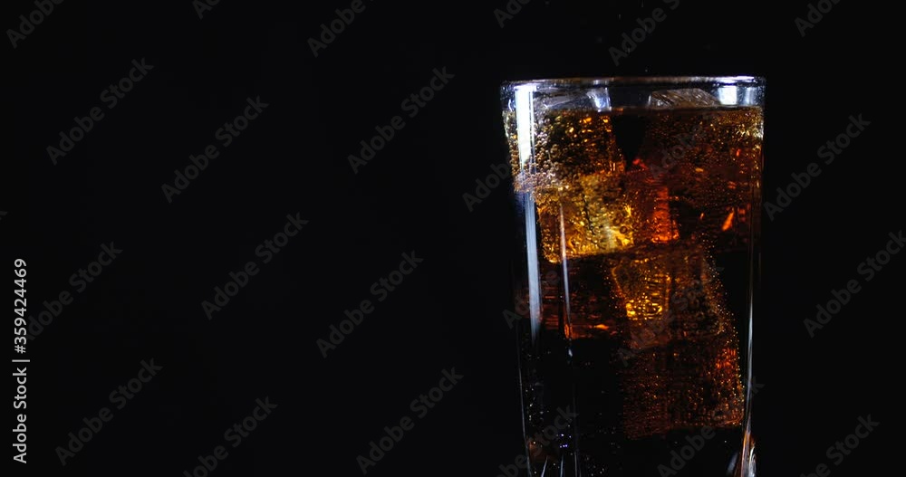 Cola with ice cubes. Side view of rotating glass in which cold carbonated brown sweet drink is poured on black background isolated with free copy space. Slow Motion 50 FPS 4k Cola with ice cubes. Side view of rotating glass in which cold carbonated brown sweet drink is poured on black background isolated with free copy space. Slow Motion 50 FPS 4k