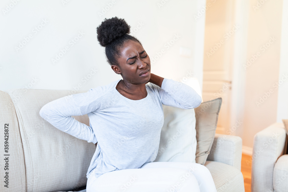 Young Black woman suffering from backache at home. Portrait of a young brunette girl sitting on the couch at home with a headache and back pain. Beautiful woman Having Spinal Or Kidney Pain Young Black woman suffering from backache at home. Portrait of a young brunette girl sitting on the couch at home with a headache and back pain. Beautiful woman Having Spinal Or Kidney Pain