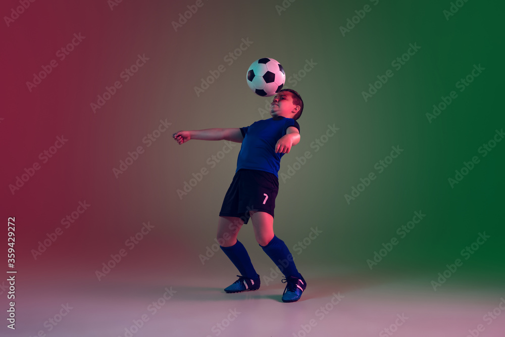 Unstoppable. Teen male football or soccer player on gradient background in neon light. Caucasian boy training, practicing on the run, in jump. Concept of sport, competition, winning, motion, action.