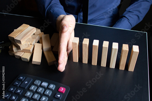 Male hand prevents wooden blocks from falling on the office desk. In the frame is a calculator, pen and coins. The concept of unemployment control, the dismissal of employees in the company 