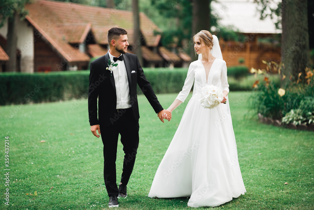 Stylish couple of happy newlyweds walking in the park on their wedding day with bouquet Stylish couple of happy newlyweds walking in the park on their wedding day with bouquet