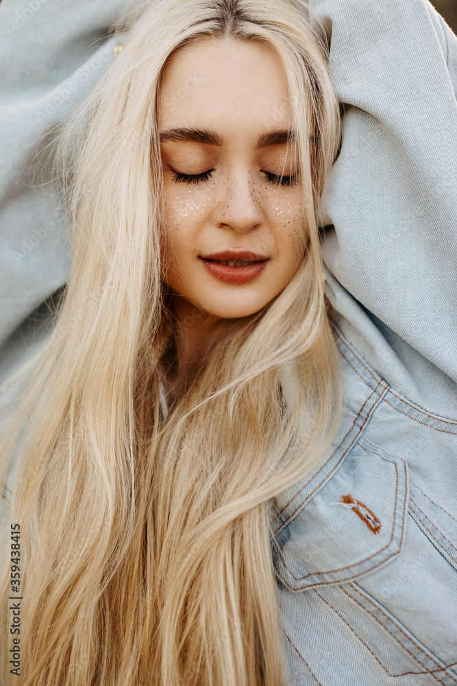 Young woman with freckles and long blonde hair, wearing a denim jacket, posing outdoors in sun light, with eyes closed. Young woman with freckles and long blonde hair, wearing a denim jacket, posing outdoors in sun light, with eyes closed.